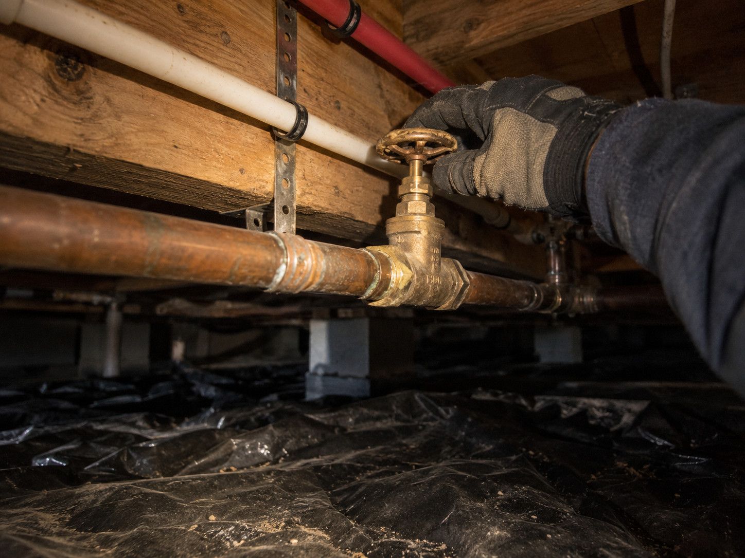 Gloved hand closing a brass gate valve on a copper water line in a dark crawl space